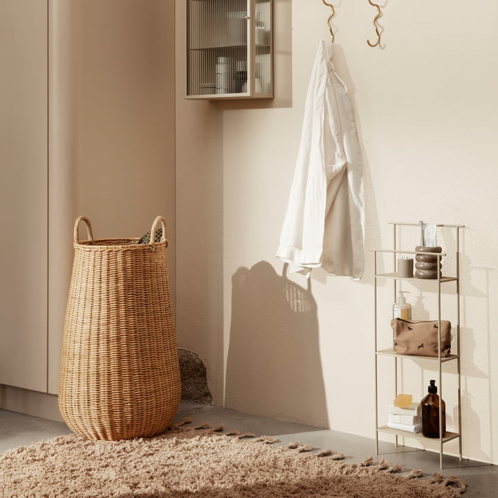 Der Braided Rattan Wäschekorb von Ferm Living auf einem beigen Teppich neben einem kleinen Regal mit Bad-Accessoires